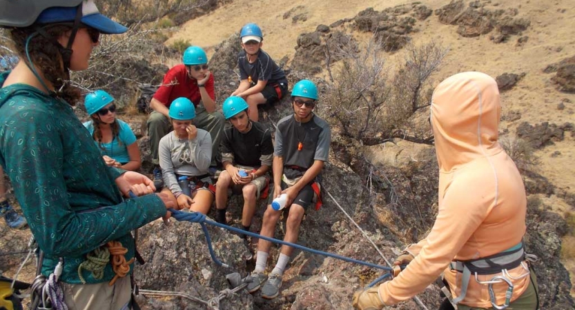 Students wearing helmets watch as an outward bound instructor provides a rock climbing lesson.